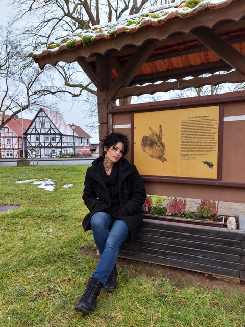AI photomontage: Mascha sits on a wooden bench beneath a covered information display in Grebendorf. A painted hare is visible on the board behind her.