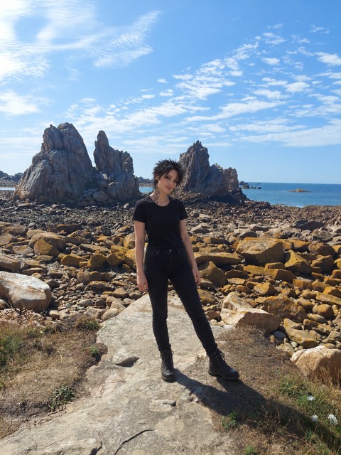 AI photomontage: Mascha stands in black clothing on a rocky coastal path in Brittany, with the sea and tall rocks behind her.