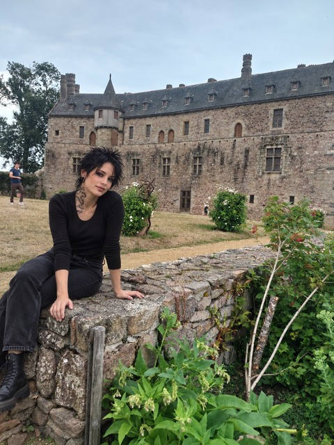 AI photomontage: Mascha sits in black clothing on a low stone wall in front of a château in Brittany, with plants growing in the foreground.