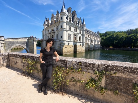 AI photomontage: Mascha leans against a stone parapet in front of Château de Chenonceau by the water.