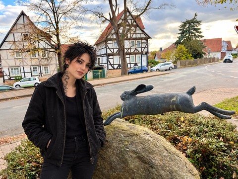 AI photomontage: Mascha stands next to a hare sculpture on a rock in Grebendorf, with half-timbered houses in the background.