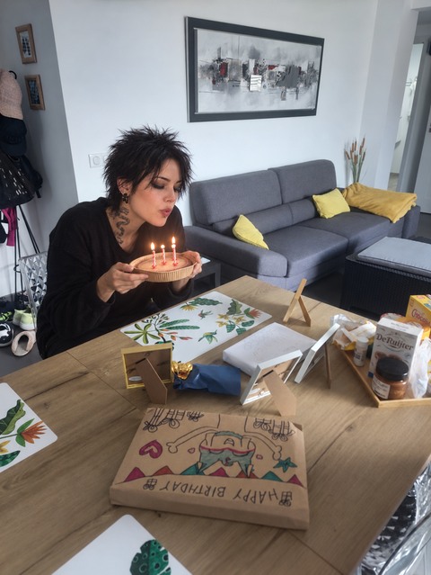 AI photomontage: At a set table, Mascha holds a small cake with three lit candles and blows toward the flames.