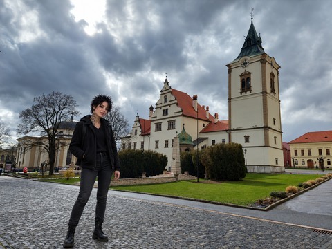 AI photomontage: Mascha stands on the left in the foreground of a paved square in Levoča, with historic buildings and a clock tower behind her.