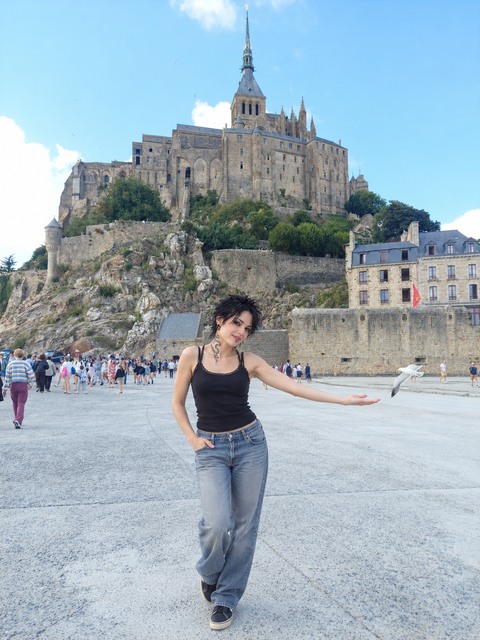 AI photomontage: At Mont-Saint-Michel, Mascha reaches one hand toward a flying seagull; the abbey and other visitors are visible behind her.