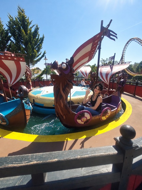 AI photomontage: Mascha sits in a dragon boat at Parc Astérix, smiling and waving.